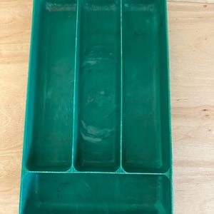 Puede incluir: Una bandeja rectangular de color verde azulado con cuatro compartimentos. La bandeja está hecha de un material sólido y duradero y está diseñada para organizar cubiertos u otras herramientas de cocina. La bandeja se encuentra sobre una superficie de madera clara.