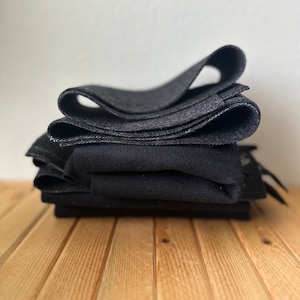 May include: A stack of folded, dark gray fabric squares. The fabric has a slightly textured appearance and is arranged on a light brown wooden surface. The folds create a layered effect, with the top layer curled.