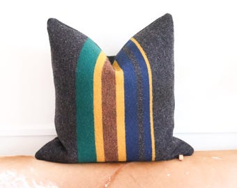 May include: A gray pillow with colorful stripes in shades of green, yellow, blue, and brown. The pillow is made of a soft, textured fabric.