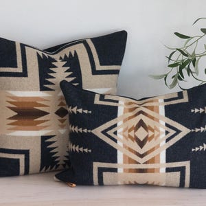 Op de afbeelding: Twee decoratieve kussens met geometrische patronen. De kussens hebben een marineblauwe achtergrond met beige, bruine en witte ontwerpen. Het grotere kussen is rechthoekig en het kleinere is vierkant.