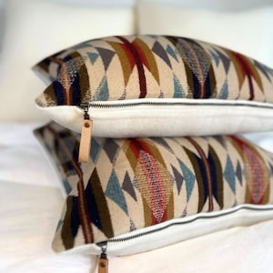 Puede incluir: Dos almohadas decorativas con un patrón geométrico en tonos marrones, azules, rojos y blancos. Las almohadas tienen una cremallera y una lengüeta de cuero.