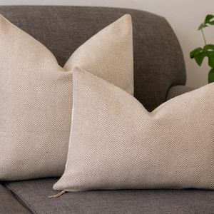 May include: Two beige throw cushions with a subtle herringbone pattern, placed on a brown sofa.