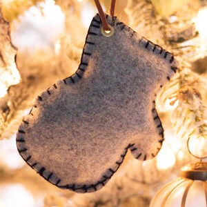 Pode incluir: Enfeite de Natal cinzento em forma de luva de feltro com costuras pretas nas bordas. O enfeite pende de um cordão de couro castanho sobre um fundo desfocado de uma árvore de Natal com luzes e uma bola decorativa.