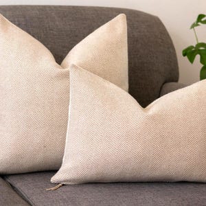 May include: Two beige decorative cushions with a herringbone pattern on a grey sofa. The cushions have a soft, textured appearance and a neutral colour palette, suitable for various interior design styles.