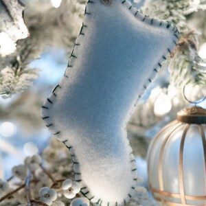 Pode incluir: Enfeite de Natal de meia de feltro azul claro com costuras verdes nas bordas. O enfeite está pendurado por um fio de cor clara contra um fundo de árvore de Natal gelada e uma bola decorativa de vidro.