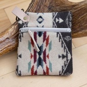 Puede incluir: Pequeño bolso rectangular con cierre de cremallera. El bolso presenta un fondo color crema con un estampado geométrico en tonos negro, rojo, verde azulado y marino. Tiene una lengüeta de cuero marrón claro y una cremallera plateada.