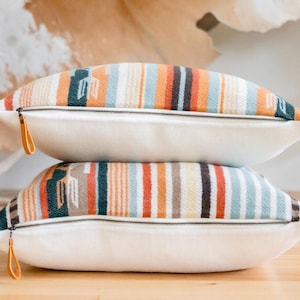 Puede incluir: Dos almohadas decorativas con un patrón de rayas coloridas. Las almohadas están apiladas una encima de la otra y tienen un respaldo blanco con una cremallera.