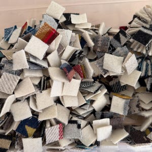 Pode incluir: Uma pilha de pequenos retalhos de tecido quadrados em várias cores e padrões. Os retalhos incluem branco, cinza, azul, vermelho e marrom, com texturas como tweed e desenhos tecidos. As peças estão contidas em um recipiente de plástico transparente.