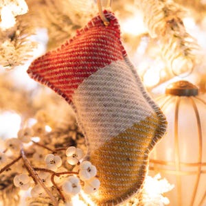 Puede incluir: Adorno navideño hecho a mano con forma de calcetín, con un diseño de bloques de color rojo, gris y amarillo. El adorno está hecho de una tela texturizada y tiene un borde cosido. Cuelga de una fina correa de cuero marrón, sobre un fondo borroso de un árbol de Navidad.