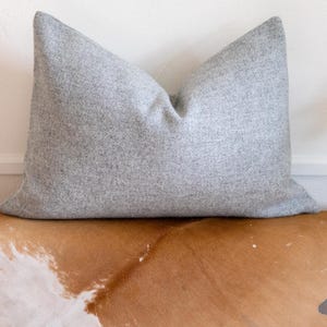 May include: A grey throw pillow with a soft, textured fabric. The pillow is sitting on a brown and white cowhide rug.