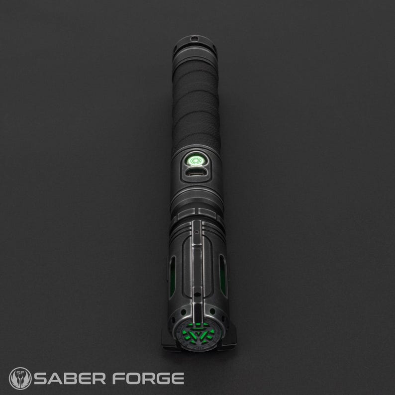 Pode incluir: Um sabre de luz preto com uma empunhadura texturizada e detalhes verdes. O punho apresenta um bot&atilde;o circular e um design detalhado com se&ccedil;&otilde;es iluminadas em verde. O logotipo Saber Forge &eacute; vis&iacute;vel.