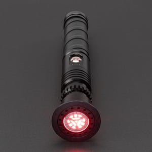 May include: A black lightsaber with a red blade. The lightsaber has a black handle with a silver band and a red button. The blade is illuminated and has a red glow.