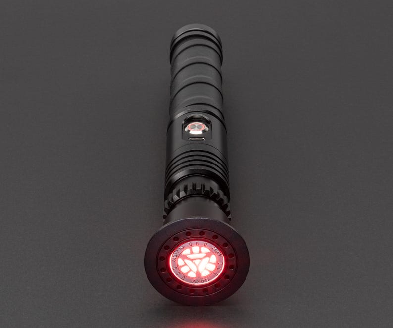 May include: A black lightsaber with a red blade. The lightsaber has a black handle with a silver band and a red button. The blade is illuminated and has a red glow.