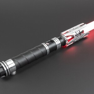 May include: A black lightsaber with a silver handle and a red blade. The lightsaber has a textured grip and a silver emitter.