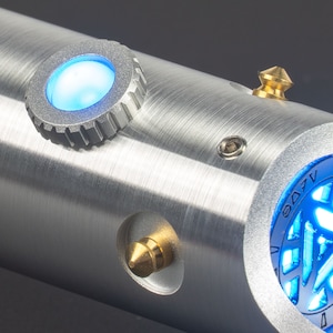 May include: A close-up of a metallic silver lightsaber hilt with blue glowing accents. The hilt features a circular blue light, a gold-colored conical button, and a circular design with blue light. The lightsaber has a brushed metal finish.