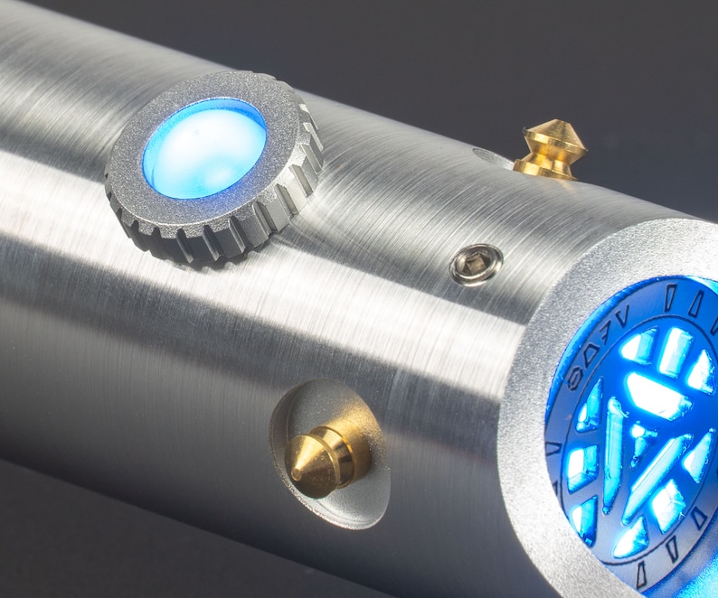 May include: A close-up of a metallic silver lightsaber hilt with blue glowing accents. The hilt features a circular blue light, a gold-colored conical button, and a circular design with blue light. The lightsaber has a brushed metal finish.