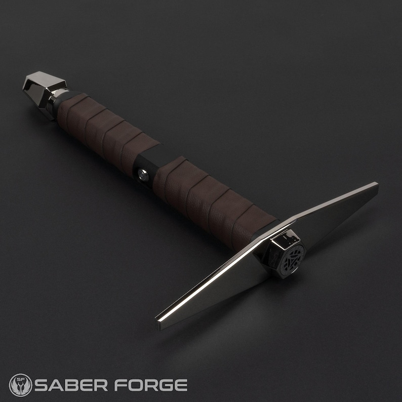 Op de afbeelding: Een zilveren en bruine lightsaber met een leren handvat en een platte, bladscherpe punt. De lightsaber is ontworpen om eruit te zien als een middeleeuws wapen.