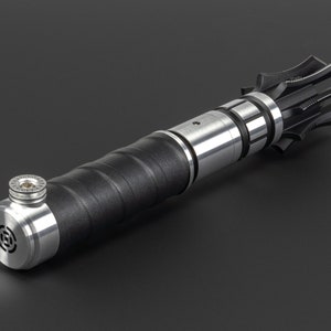 May include: A black and silver lightsaber prop with a textured black handle and a silver blade holder. The blade holder has a silver ring with a small screw in the centre.