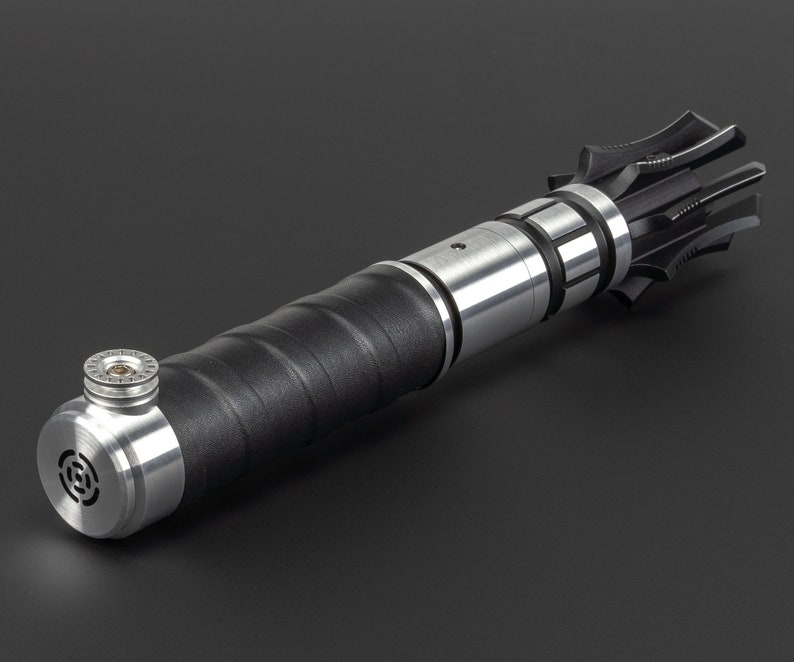 May include: A black and silver lightsaber prop with a textured black handle and a silver blade holder. The blade holder has a silver ring with a small screw in the centre.