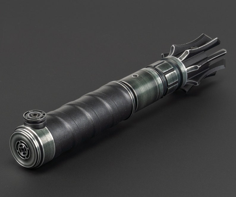 K&ouml;nnte beinhalten: Ein schwarzes und gr&uuml;nes Lichtschwert mit einem detaillierten, komplizierten Design. Das Lichtschwert hat einen strukturierten Griff und einen silberfarbenen Emitter.