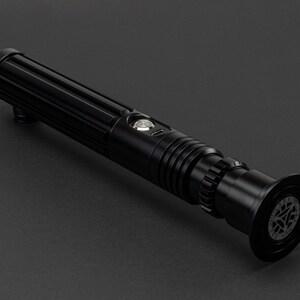 Op de afbeelding: Een zwart lightsaber met een getextureerde handgreep en een ronde emitter met een zilveren knop. De emitter heeft een zwart en zilver ontwerp met een rond logo.