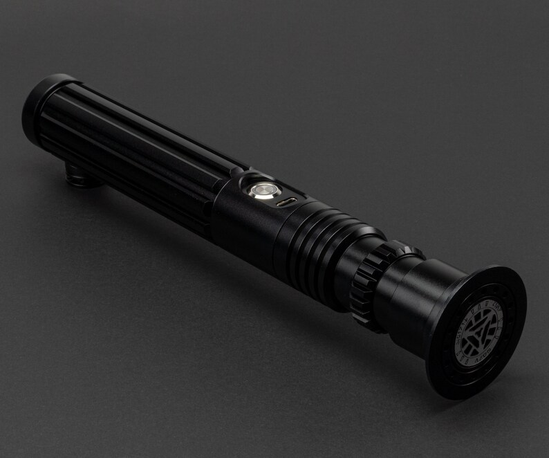 Op de afbeelding: Een zwart lightsaber met een getextureerde handgreep en een ronde emitter met een zilveren knop. De emitter heeft een zwart en zilver ontwerp met een rond logo.