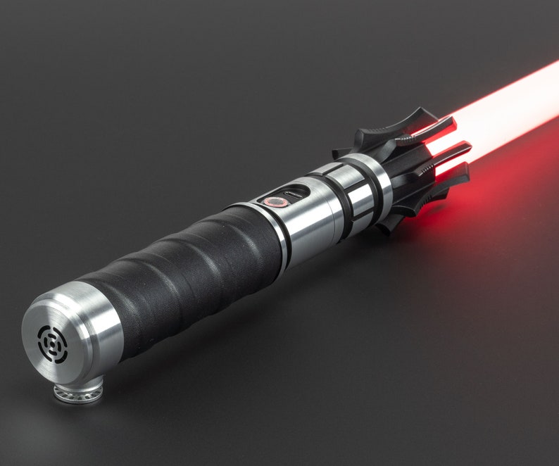 May include: A silver and black lightsaber with a red blade. The lightsaber has a textured black grip and a silver emitter with a red button.