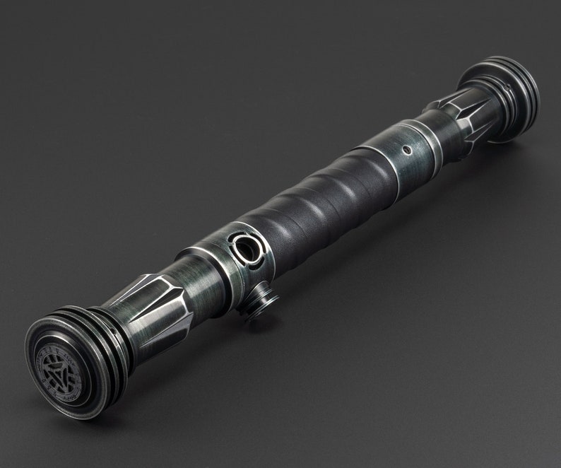 Pode incluir: Um sabre de luz preto e prateado com uma pega texturizada e um emissor circular. O sabre de luz tem um acabamento met&aacute;lico verde escuro.