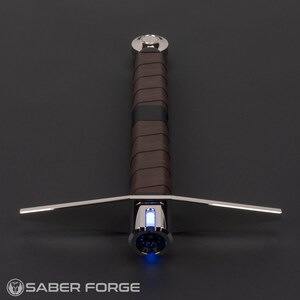 May include: A silver-toned metal hilt with a brown leather-wrapped grip and a crossguard. The hilt features a glowing blue light at the emitter and a polished pommel at the base. The Saber Forge logo is visible in the bottom left corner.