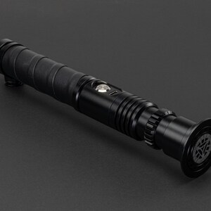 May include: A black lightsaber with a textured grip and a silver button. The lightsaber has a black emitter with a circular design.