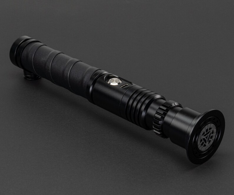 May include: A black lightsaber with a textured grip and a silver button. The lightsaber has a black emitter with a circular design.