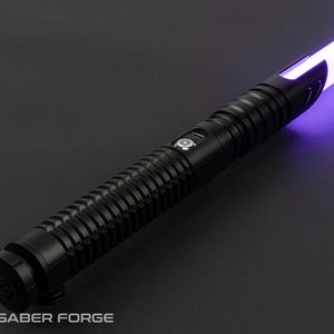 May include: A black lightsaber with a ribbed handle and a glowing purple blade. The hilt features a button and a USB port. The Saber Forge logo is visible at the base of the hilt.