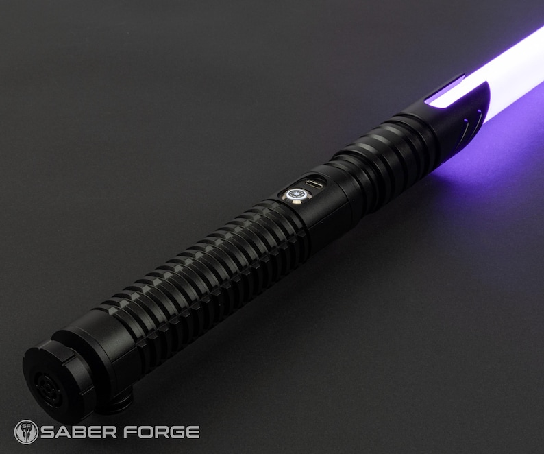 May include: A black lightsaber with a ribbed handle and a glowing purple blade. The hilt features a button and a USB port. The Saber Forge logo is visible at the base of the hilt.