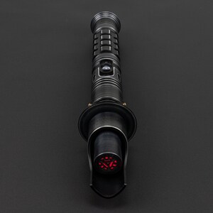 K&ouml;nnte beinhalten: Ein schwarzes Lichtschwert mit einer roten Klinge. Das Lichtschwert hat einen schwarzen Griff mit einem silbernen Knopf und einen schwarzen Emitter.