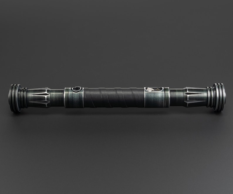 Pode incluir: Um sabre de luz preto com acabamento verde e prateado. O sabre de luz tem uma pega texturizada e um bot&atilde;o prateado no lado.