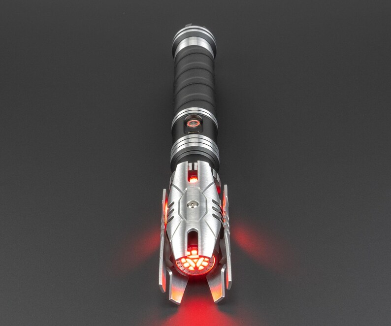 May include: A black and silver lightsaber with a red blade. The lightsaber has a detailed design with a textured grip and a silver emitter.