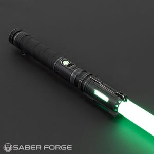 Pode incluir: Um sabre de luz preto com uma l&acirc;mina verde brilhante. O punho &eacute; preto com detalhes prateados e uma pega texturizada. O sabre de luz tem um bot&atilde;o com luz verde e o logotipo Saber Forge.