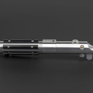 May include: A silver lightsaber with black accents and a curved emitter. The hilt features a ribbed design with black grips and various buttons and knobs. The lightsaber is a prop from a science fiction franchise.