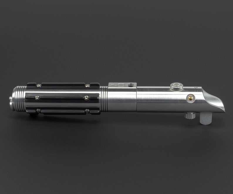 May include: A silver lightsaber with black accents and a curved emitter. The hilt features a ribbed design with black grips and various buttons and knobs. The lightsaber is a prop from a science fiction franchise.