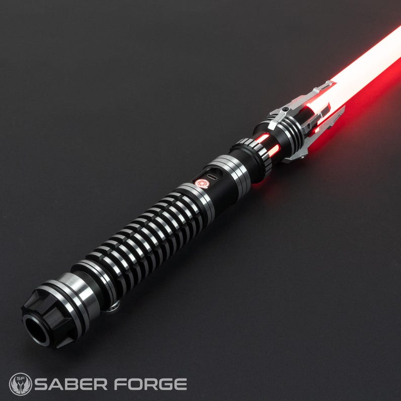 Puede incluir: Un sable de luz negro y plateado con una hoja roja. El sable de luz tiene un agarre texturizado y un emisor plateado. La hoja est&aacute; encendida y se extiende desde el emisor.