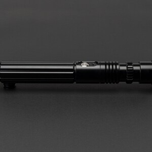 Op de afbeelding: Een zwart metalen lightsaber-accessoire met een getextureerde handgreep en een zilveren knop. De lightsaber is ongeveer 30 cm lang.