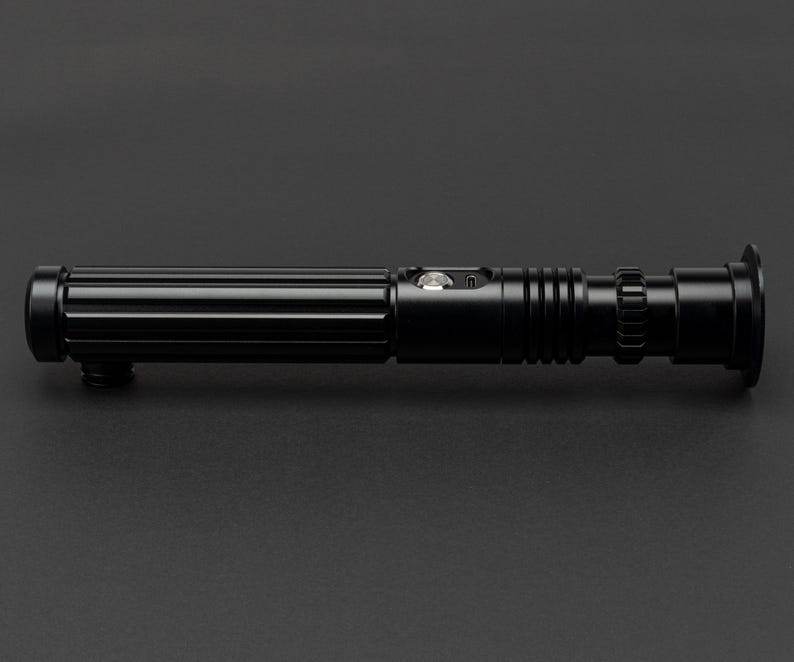 Op de afbeelding: Een zwart metalen lightsaber-accessoire met een getextureerde handgreep en een zilveren knop. De lightsaber is ongeveer 30 cm lang.