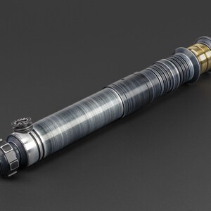 Puede incluir: Un sable de luz plateado y negro con un acento dorado en el extremo. El sable de luz tiene una superficie texturizada y un dise&ntilde;o detallado.