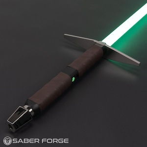 Op de afbeelding: Een groene lightsaber met een bruine leren handvat en een zilveren metalen handvat. De lightsaber is aan en de blade gloeit groen. De lightsaber ligt op een zwart oppervlak. SABER FORGE is afgedrukt op het oppervlak.