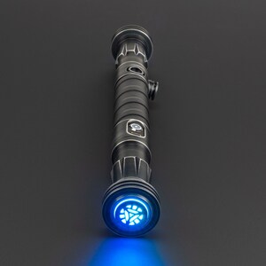 Pode incluir: Um sabre de luz preto com uma l&acirc;mina de sabre de luz azul e um detalhe prateado no cabo. O sabre de luz est&aacute; ligado e a luz azul ilumina a superf&iacute;cie abaixo.