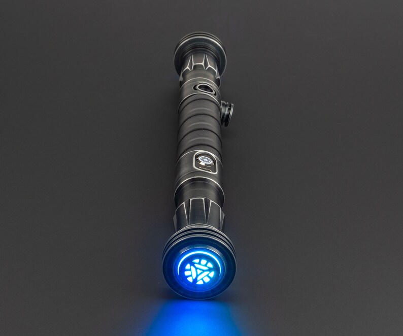 Pode incluir: Um sabre de luz preto com uma l&acirc;mina de sabre de luz azul e um detalhe prateado no cabo. O sabre de luz est&aacute; ligado e a luz azul ilumina a superf&iacute;cie abaixo.