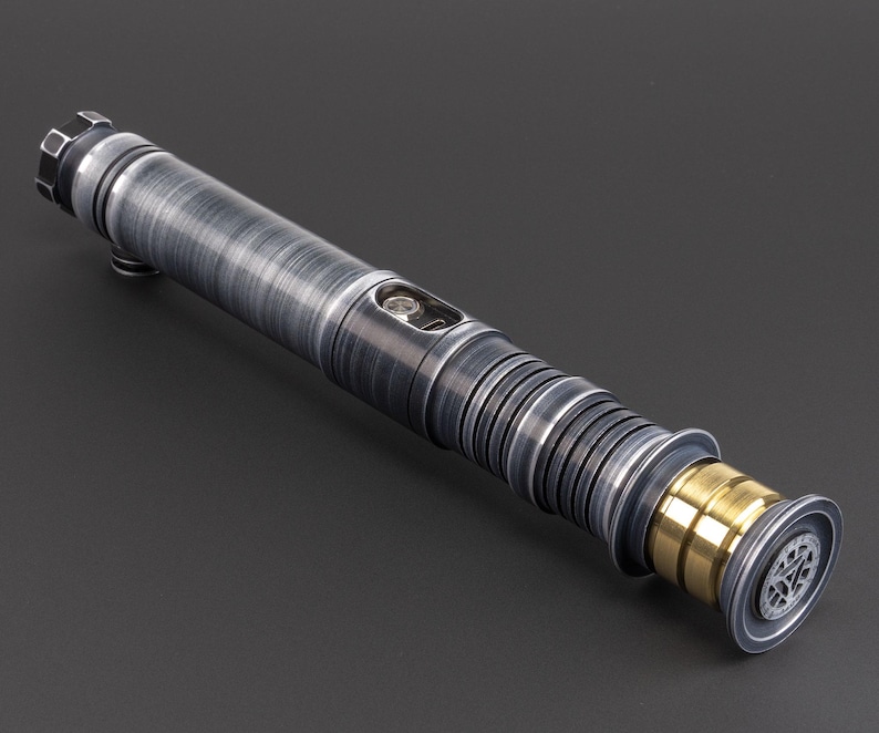 Puede incluir: Un sable de luz plateado y negro con un acento dorado. El sable de luz tiene una superficie texturizada y un dise&ntilde;o detallado.