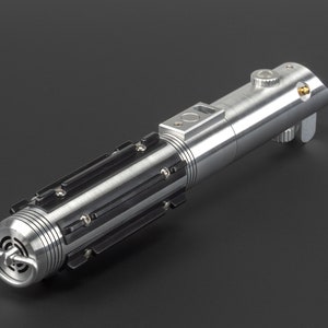 May include: A metallic silver lightsaber with black accents. The hilt features a cylindrical design with detailed ridges, panels, and a circular emitter. A button and a small gold-colored detail are visible on the top. The lightsaber is a prop or collectible.