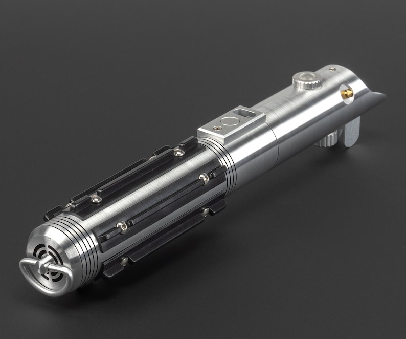 May include: A metallic silver lightsaber with black accents. The hilt features a cylindrical design with detailed ridges, panels, and a circular emitter. A button and a small gold-colored detail are visible on the top. The lightsaber is a prop or collectible.