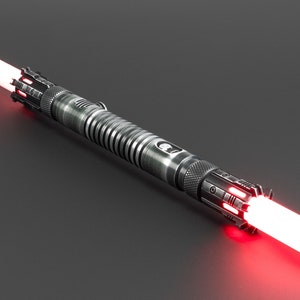 Op de afbeelding: Een dubbelzijdige lichtzwaard met een metallic zilveren afwerking. Het handvat heeft ingewikkelde details en een geribbeld middengedeelte. Elk uiteinde straalt een helder rood blad uit. Het ontwerp is futuristisch en strak.